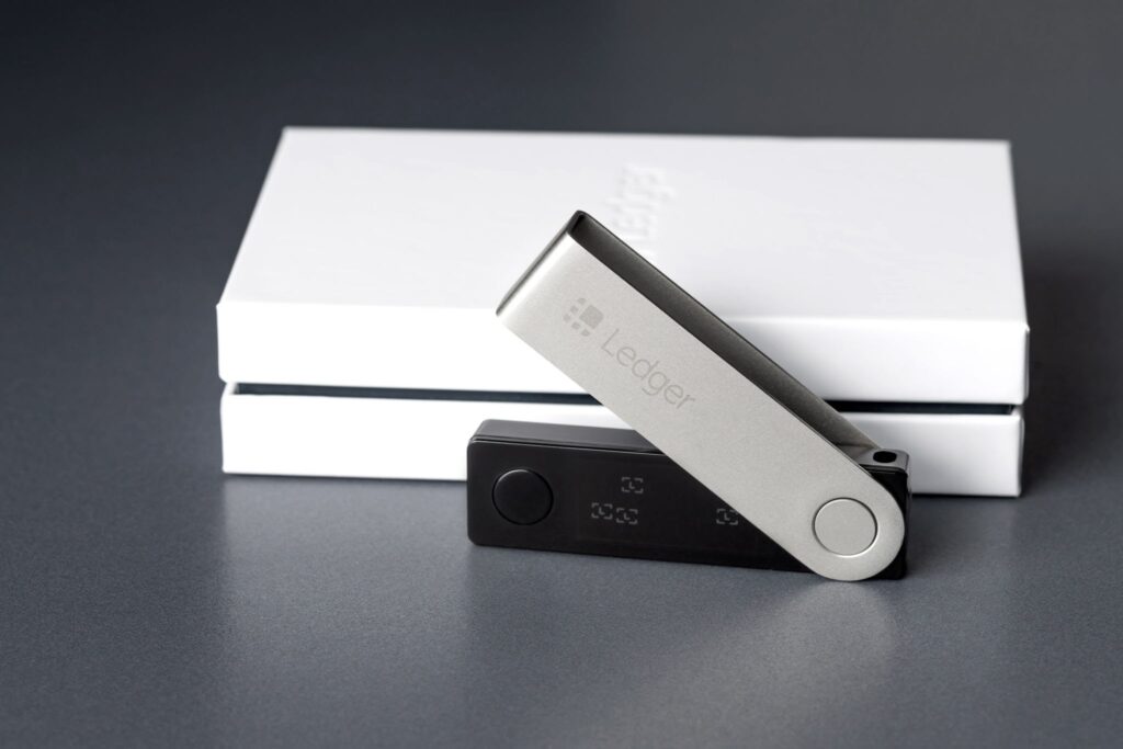 Ledger Nano X hardware wallet shown alongside its packaging and a cryptocurrency display on a computer screen.